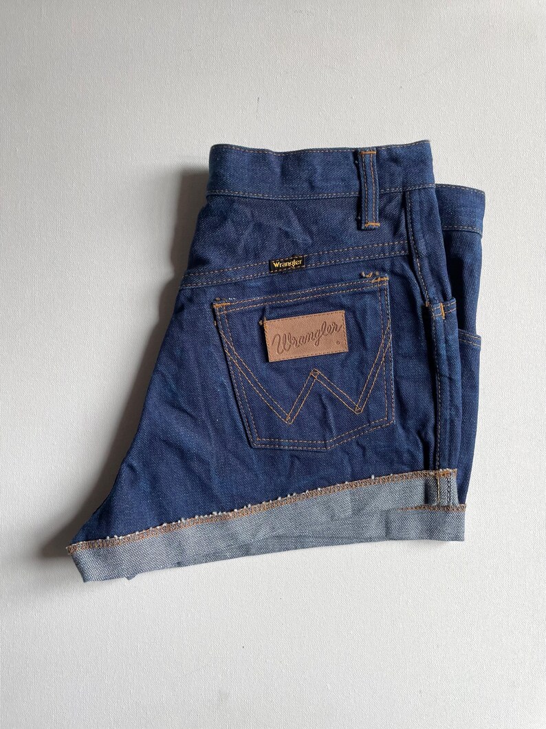 May include: Dark blue denim Wrangler shorts with a folded hem. The shorts feature the Wrangler logo on the back pocket and a small logo above the back pocket. The stitching is a contrasting tan color.