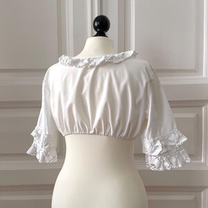 May include: A white, cropped blouse with a ruffled neckline and sleeves. The sleeves feature lace trim at the cuffs. The blouse is displayed on a mannequin against a white background.