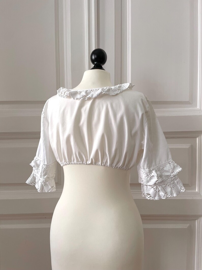 May include: A white, cropped blouse with a ruffled neckline and sleeves. The sleeves feature lace trim at the cuffs. The blouse is displayed on a mannequin against a white background.