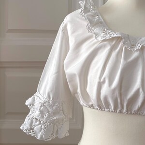 May include: White crop top with a square neckline and three-quarter sleeves. The neckline and sleeve cuffs feature delicate, openwork lace trim. The top is displayed on a mannequin.