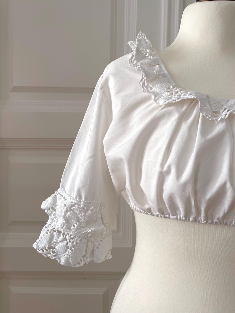 May include: White crop top with a square neckline and three-quarter sleeves. The neckline and sleeve cuffs feature delicate, openwork lace trim. The top is displayed on a mannequin.