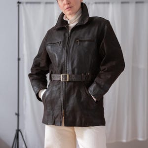 May include: A dark brown leather jacket with a belted waist and a zip-up front. The jacket has a collar and two chest pockets. The person is wearing a white turtleneck and cream-coloured trousers.