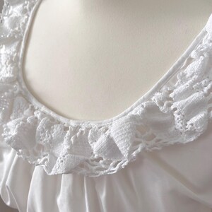 May include: Close-up of a white, vintage-style blouse. The blouse features a square neckline and a delicate, crocheted lace trim around the neckline. The fabric appears to be a soft, flowing material.