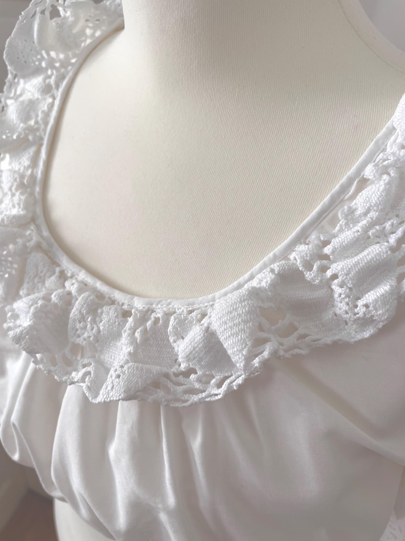 May include: Close-up of a white, vintage-style blouse. The blouse features a square neckline and a delicate, crocheted lace trim around the neckline. The fabric appears to be a soft, flowing material.