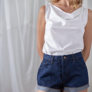 May include: A white sleeveless top with a cowl neckline paired with dark blue denim shorts. The shorts have a classic five-pocket design and rolled-up cuffs. The outfit is casual and suitable for warm weather.