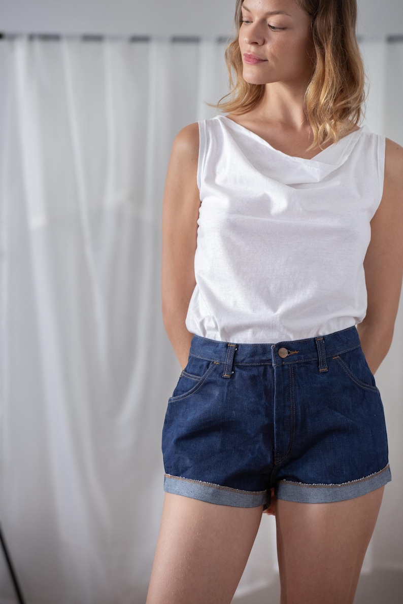 May include: A white sleeveless top with a cowl neckline paired with dark blue denim shorts. The shorts have a classic five-pocket design and rolled-up cuffs. The outfit is casual and suitable for warm weather.
