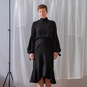 Pode incluir: Uma mulher vestindo uma blusa preta de gola alta com mangas compridas e bufantes e uma saia preta assim&eacute;trica. Ela est&aacute; em p&eacute; na frente de um fundo de cortina branca.