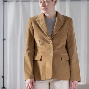 May include: A tan corduroy blazer with a two-button fastening and two front pockets. The blazer is worn over a grey t-shirt and paired with off-white trousers. The background features a white curtain and a photography stand.