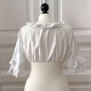 May include: A white, cropped blouse with a ruffled neckline and short sleeves. The sleeves feature lace detailing at the cuffs. The blouse is displayed on a mannequin against a white background.