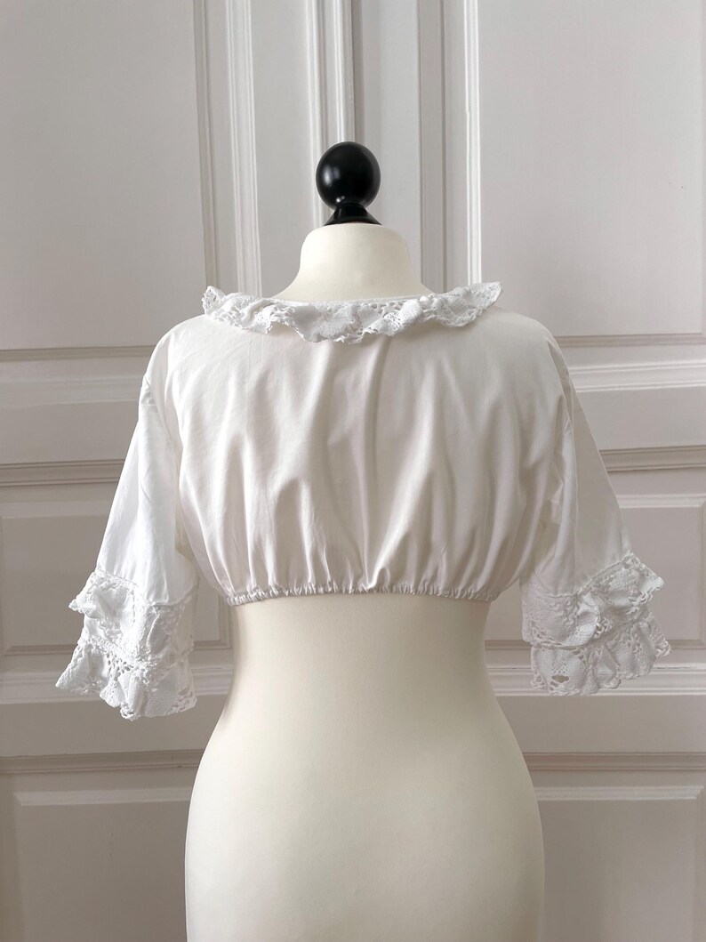 May include: A white, cropped blouse with a ruffled neckline and short sleeves. The sleeves feature lace detailing at the cuffs. The blouse is displayed on a mannequin against a white background.
