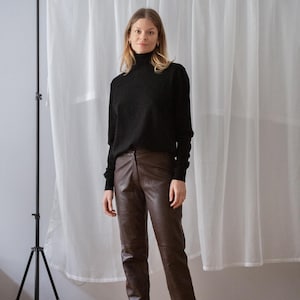 May include: A person wearing a black turtleneck sweater and brown leather pants. The pants are a dark brown color and have a straight leg fit. The background is a white curtain and a black tripod.