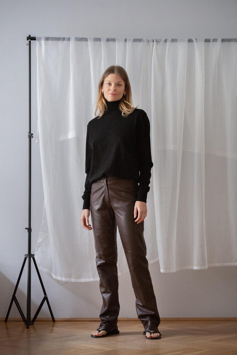 May include: A person wearing a black turtleneck sweater and brown leather pants. The pants are a dark brown color and have a straight leg fit. The background is a white curtain and a black tripod.