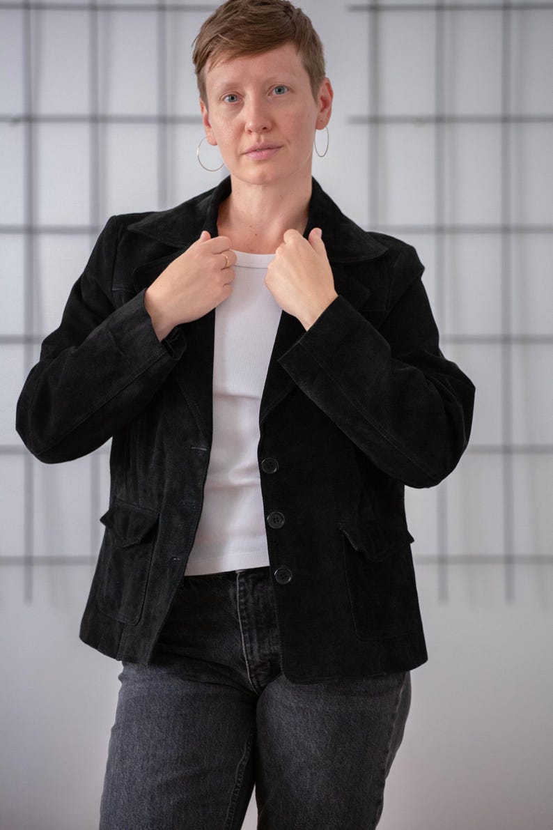 May include: A black suede jacket with a collared neckline, button closure, and two flap pockets. The jacket is being worn by a person with short brown hair.