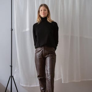 May include: A person wearing a black turtleneck sweater and brown leather pants. The pants are a dark brown color and have a straight leg design. The person is standing in front of a white curtain.