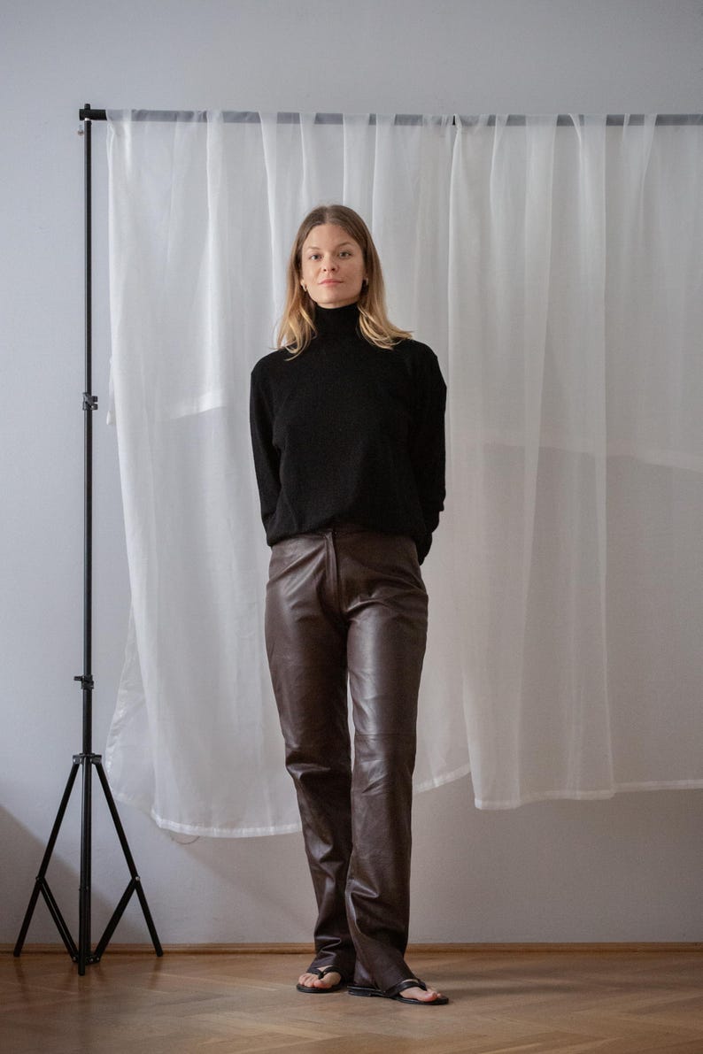May include: A person wearing a black turtleneck sweater and brown leather pants. The pants are a dark brown color and have a straight leg design. The person is standing in front of a white curtain.