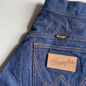 May include: Close-up of the back pocket of blue denim Wrangler jeans. The pocket has a tan leather patch with the Wrangler logo. The stitching is a contrasting tan color. The Wrangler label is visible at the waistband.