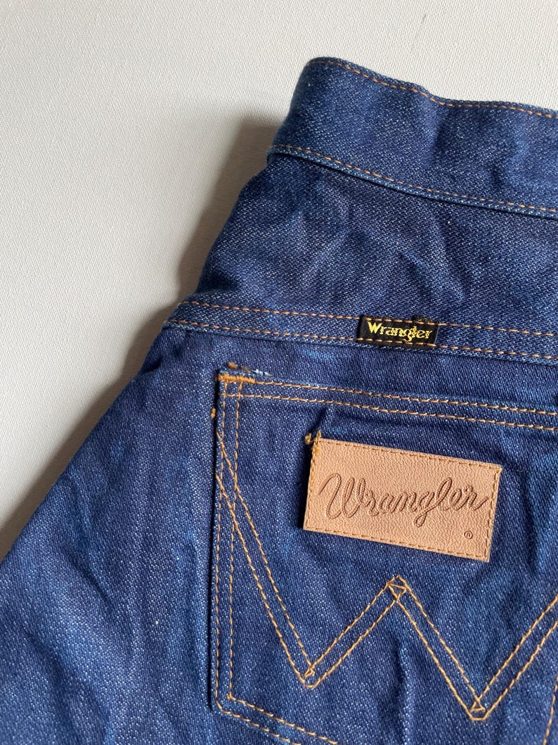 May include: Close-up of the back pocket of blue denim Wrangler jeans. The pocket has a tan leather patch with the Wrangler logo. The stitching is a contrasting tan color. The Wrangler label is visible at the waistband.