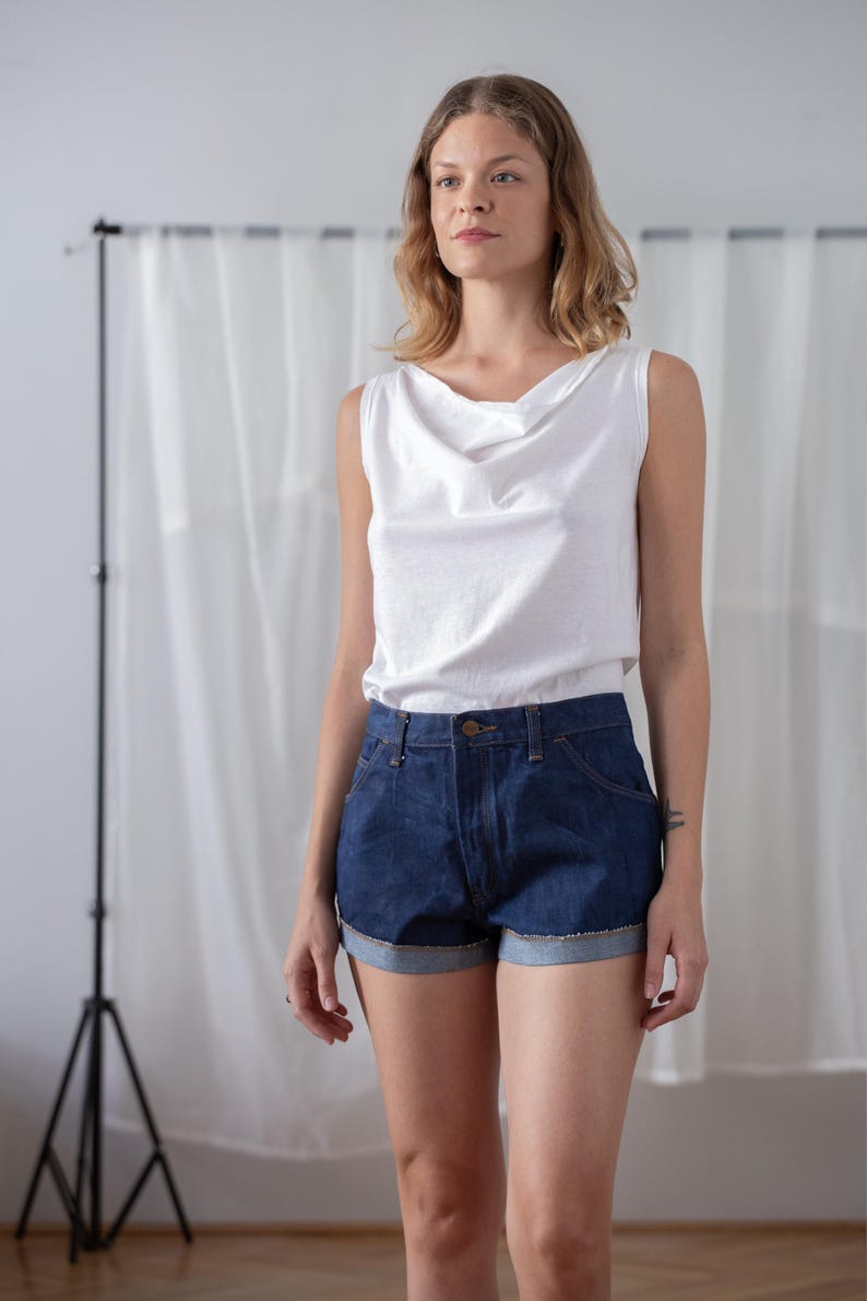 May include: A white cowl neck tank top paired with dark wash denim shorts. The shorts have a rolled hem and classic five-pocket design. The outfit is simple and casual, suitable for warm weather.