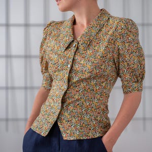 Può includere: Una donna indossa una blusa a maniche corte con stampa floreale, colletto e chiusura con bottoni. Indossa anche pantaloni blu scuro.