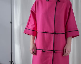 May include: A long, pink coat with black trim and a black belt. The coat has a unique design with a diagonal seam and multiple black buckles.