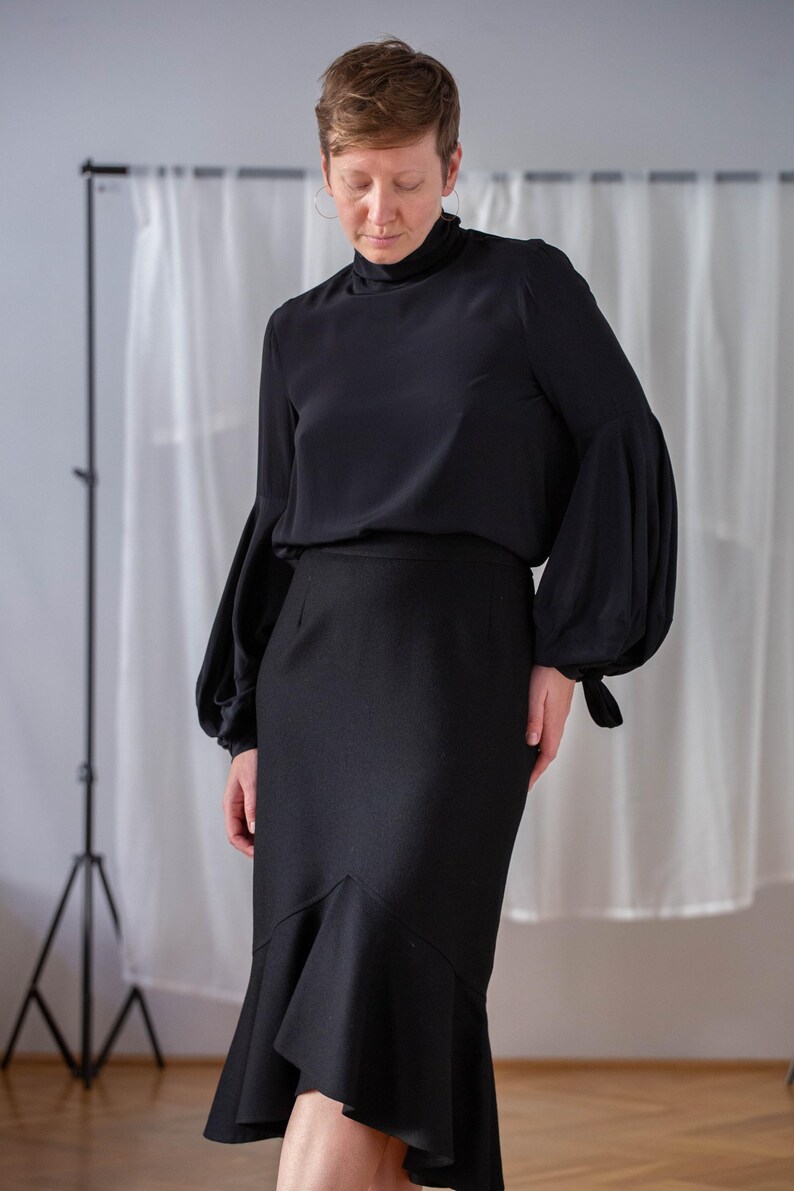 Pode incluir: Uma blusa preta de gola alta com mangas compridas e volumosas, combinada com uma saia midi preta com uma bainha franjada.