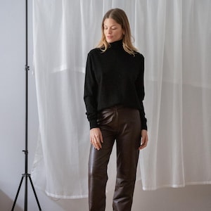 May include: A black turtleneck sweater paired with brown leather pants. The pants have a slight flare at the bottom. The outfit is completed with black sandals. The background features a white curtain and a black tripod.