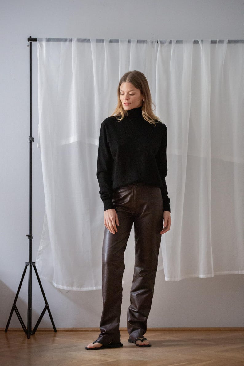 May include: A black turtleneck sweater paired with brown leather pants. The pants have a slight flare at the bottom. The outfit is completed with black sandals. The background features a white curtain and a black tripod.