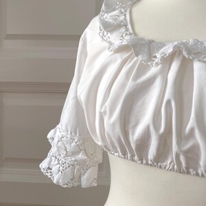 May include: White crop top with a ruffled neckline and short sleeves. The sleeves and neckline are trimmed with delicate white lace. The top is displayed on a mannequin, showcasing its design and texture. The fabric appears soft and slightly gathered.