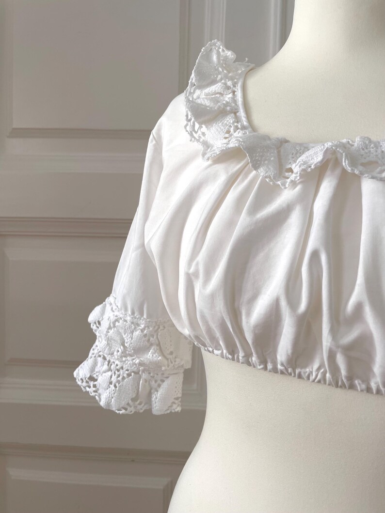 May include: White crop top with a ruffled neckline and short sleeves. The sleeves and neckline are trimmed with delicate white lace. The top is displayed on a mannequin, showcasing its design and texture. The fabric appears soft and slightly gathered.