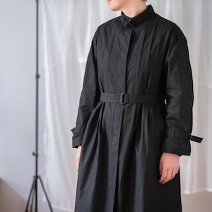 Può includere: Lungo trench nero con cintura in vita e polsini abbottonati. Il cappotto presenta un colletto alto e pieghe verticali sul corpetto. Il tessuto sembra leggero, adatto alla mezza stagione. Misura circa 130 cm di lunghezza.