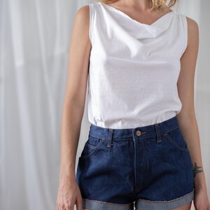 May include: A white sleeveless top with a cowl neck paired with dark blue denim shorts. The shorts have a rolled hem and classic five-pocket styling. The outfit is casual and suitable for warm weather.