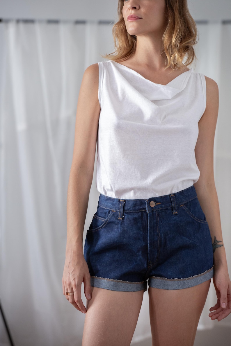 May include: A white sleeveless top with a cowl neck paired with dark blue denim shorts. The shorts have a rolled hem and classic five-pocket styling. The outfit is casual and suitable for warm weather.