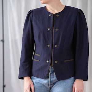 May include: A navy blue jacket with a round neckline and gold buttons. The jacket has a tailored fit with subtle shoulder pads and brown trim on the pockets. The sleeves are long and the jacket is worn with blue jeans.