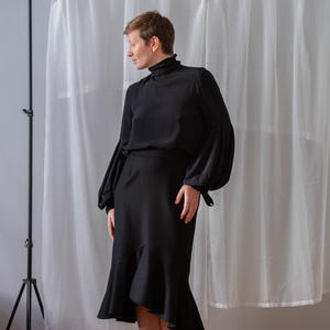 Pode incluir: Uma mulher vestindo uma blusa preta de gola alta com mangas compridas e uma saia preta com bainha franjada. A saia &eacute; assim&eacute;trica, com um lado mais longo que o outro. Ela est&aacute; em p&eacute; na frente de uma cortina branca.