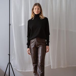 May include: A woman wearing a black turtleneck sweater and brown leather pants. The pants have a straight leg design. The background includes a white curtain and a black tripod.