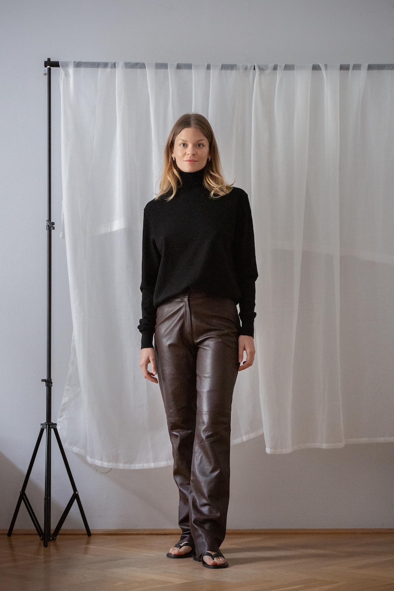 May include: A woman wearing a black turtleneck sweater and brown leather pants. The pants have a straight leg design. The background includes a white curtain and a black tripod.