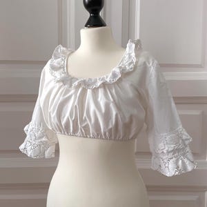 May include: White crop top with a ruffled neckline and sleeves. The sleeves are three-quarter length and feature lace trim at the cuffs. The top is displayed on a mannequin.