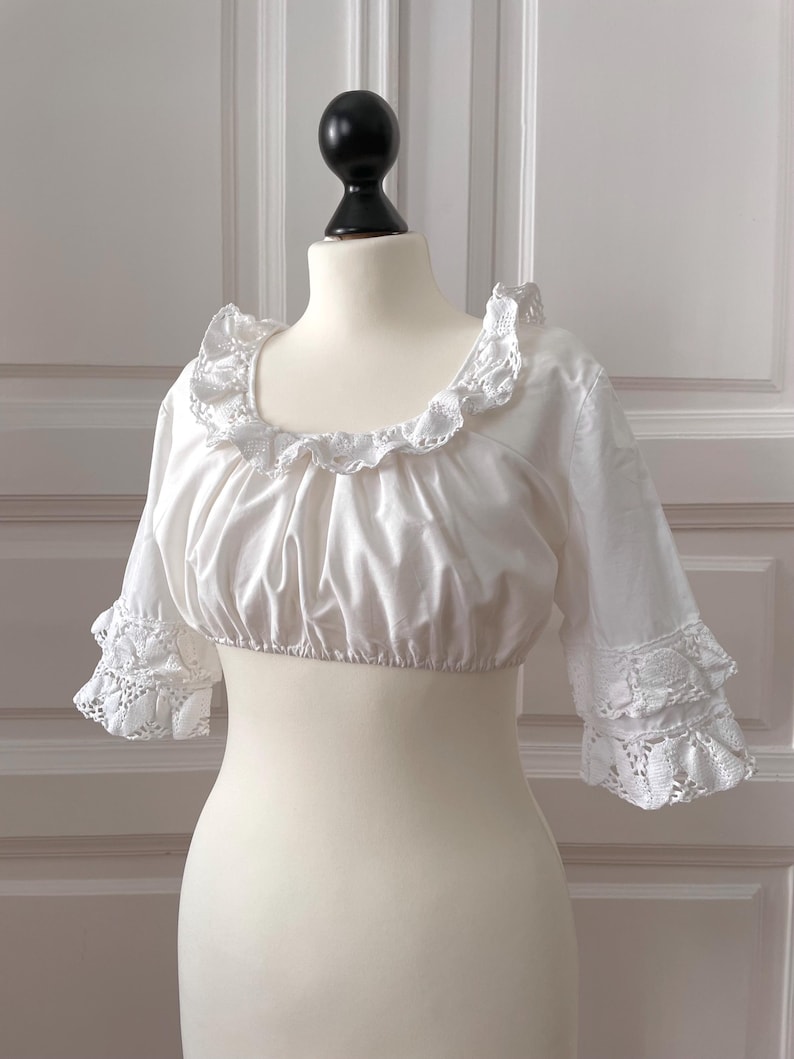 May include: White crop top with a ruffled neckline and sleeves. The sleeves are three-quarter length and feature lace trim at the cuffs. The top is displayed on a mannequin.