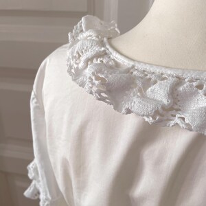 May include: A white blouse with a detailed, crocheted lace collar and sleeve trim. The close-up shot highlights the texture and delicate design of the lace against the smooth fabric of the garment. The blouse is displayed on a mannequin.
