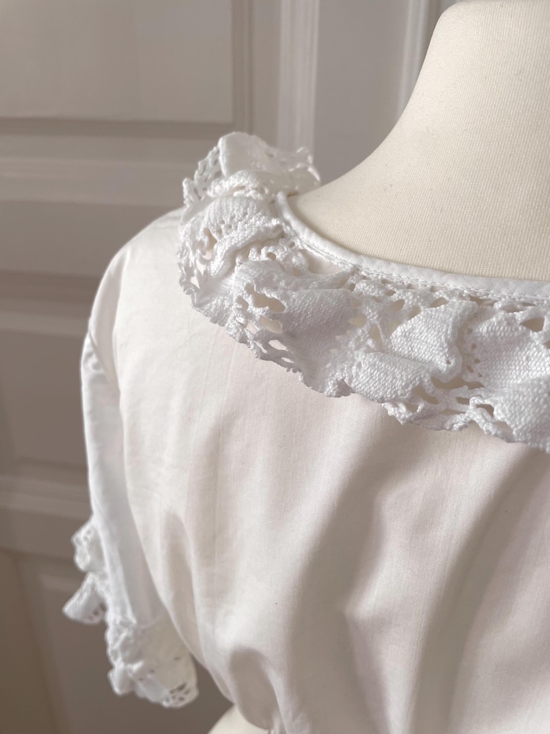 May include: A white blouse with a detailed, crocheted lace collar and sleeve trim. The close-up shot highlights the texture and delicate design of the lace against the smooth fabric of the garment. The blouse is displayed on a mannequin.