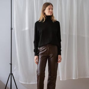 May include: A woman wearing a black turtleneck sweater and brown leather pants. The pants are a rich, dark brown color. The outfit is paired with black sandals. The background includes a white curtain and a black tripod.