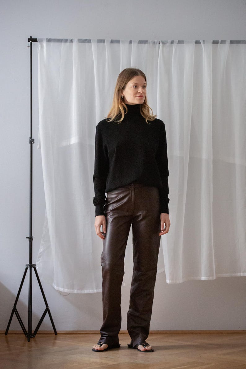 May include: A woman wearing a black turtleneck sweater and brown leather pants. The pants are a rich, dark brown color. The outfit is paired with black sandals. The background includes a white curtain and a black tripod.