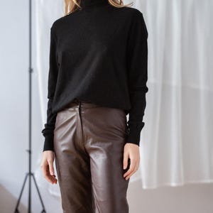 May include: A black turtleneck sweater paired with brown leather pants. The sweater has long sleeves and a high neckline. The pants are a rich brown color and have a classic, tailored fit. The outfit is simple and elegant.