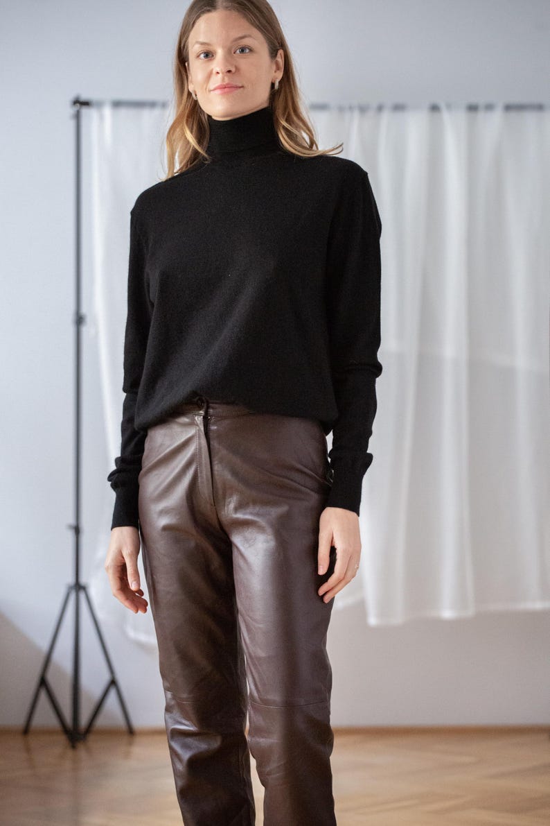 May include: A black turtleneck sweater paired with brown leather pants. The sweater has long sleeves and a high neckline. The pants are a rich brown color and have a classic, tailored fit. The outfit is simple and elegant.