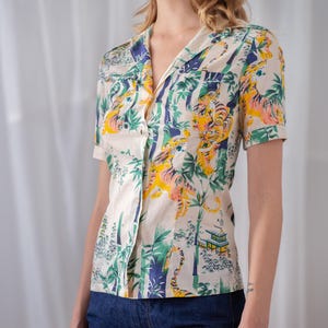 May include: Cream-colored button-up shirt with a vibrant tiger and bamboo print. The short-sleeved shirt features a classic collar and two small chest pockets. The design includes yellow, orange, green, and blue elements.