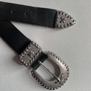 Könnte beinhalten: Schwarzer Ledergürtel mit silberfarbener Metallschnalle und Spitze. Die Schnalle hat eine abgerundete Form mit dekorativen Nähten am Rand. Die Gürtelspitze hat ein ähnliches Design. Auf weißem Hintergrund.