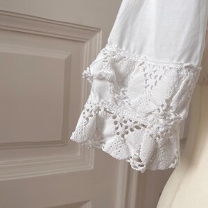 May include: Close-up of a white long-sleeve shirt sleeve with intricate lace detailing at the cuff. The cuff features a layered design with delicate openwork patterns, creating a vintage or romantic aesthetic.