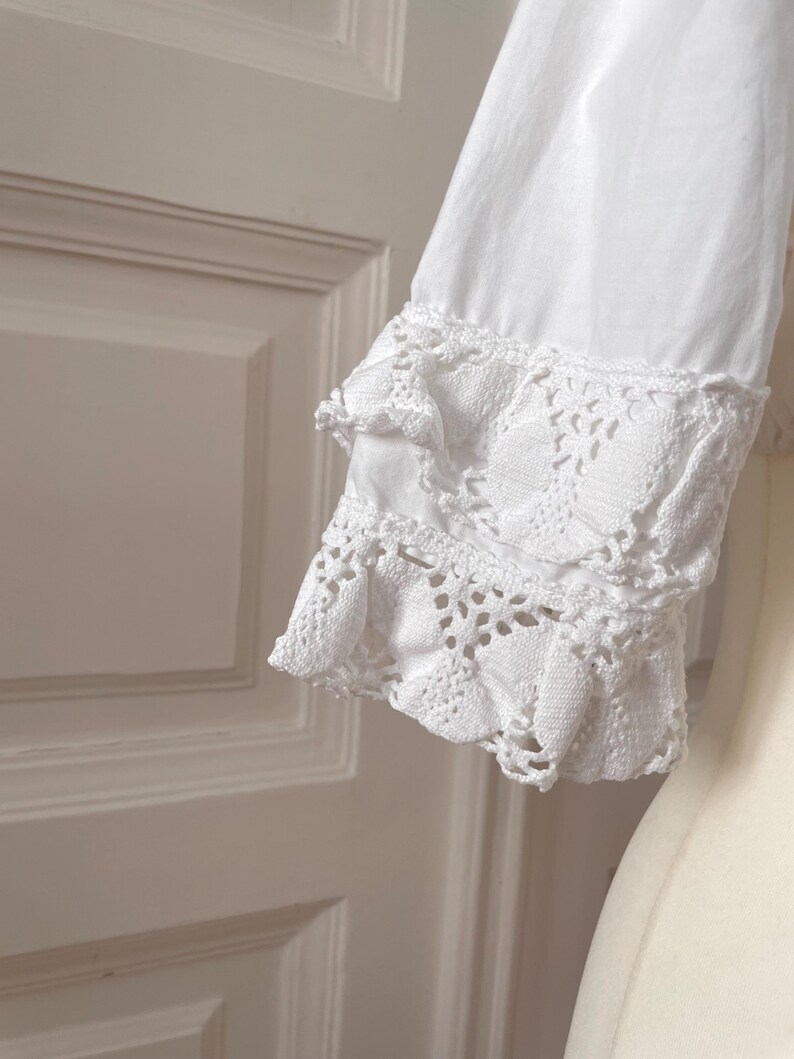 May include: Close-up of a white long-sleeve shirt sleeve with intricate lace detailing at the cuff. The cuff features a layered design with delicate openwork patterns, creating a vintage or romantic aesthetic.