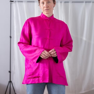May include: A vibrant hot pink long-sleeved jacket featuring a mandarin collar, traditional frog closures down the front, and a subtle tonal circular pattern throughout the fabric. The jacket has a straight hemline and two front patch pockets.