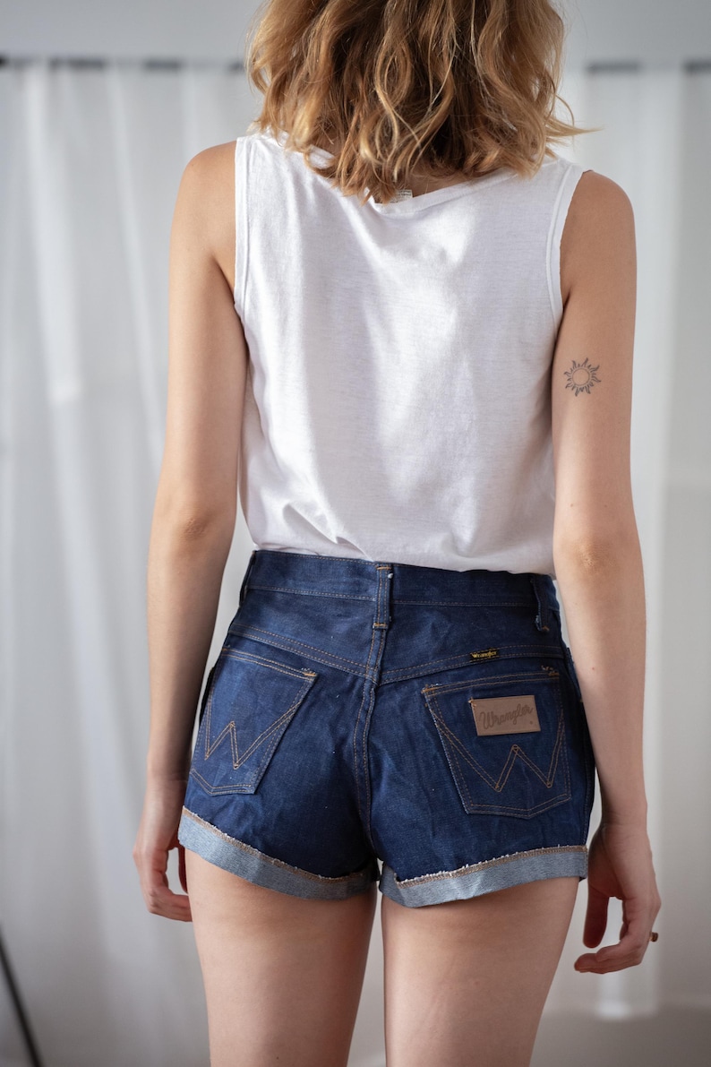 May include: Dark wash denim Wrangler shorts with a rolled hem and a brown leather patch. The shorts have a high waist and classic five-pocket styling. A white tank top is also visible.